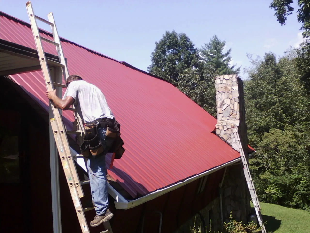 Roofer on a ladder beside a red metal roof and stone chimney — Soffit & Fascia Repair by Roofing Brothers in Fuquay Varina